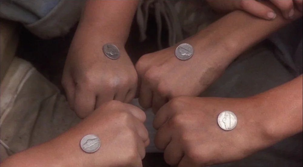 Stand By Me (1986) movie still: closeup — Four fists are arranged in a circular formation on a patch of dirt ground. Each fist has a single US…