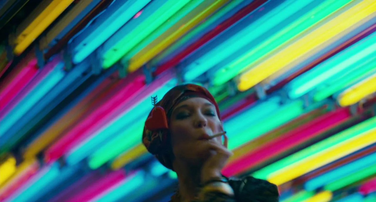 Saint Laurent (2014) movie still: closeup — A dense array of parallel, multi-colored neon light tubes stretches across the ceiling. A woman is…
