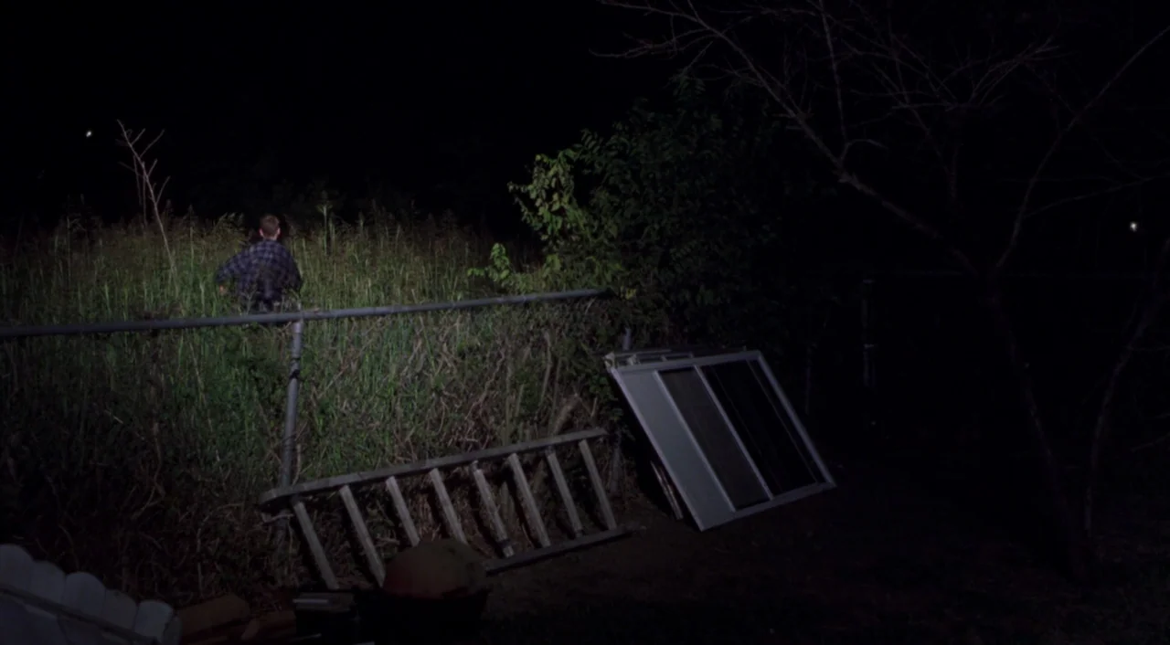 Boys Don’t Cry (1999) movie still: wide — A man climbs over a wire fence at night into a patch of tall weeds. A discarded ladder and a sliding…
