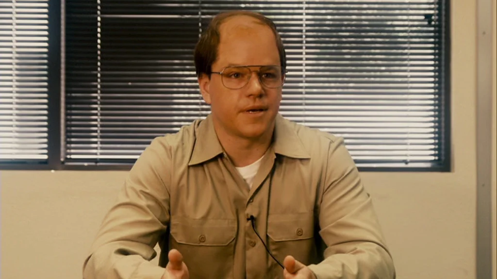 The Informant! (2009) movie still: medium — A man sits in front of black horizontal window blinds inside a sterile room. He talks while wearing a…