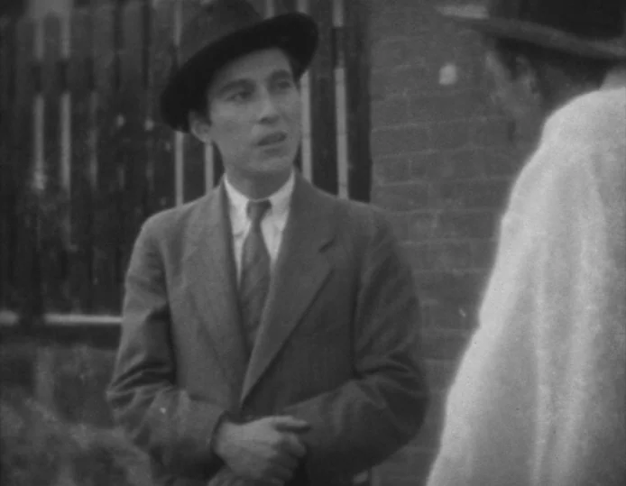 Tokyo Chorus (1931) movie still: medium — An outdoor setting features a dark wooden picket fence and a brick wall. A man in a dark suit speaks to…