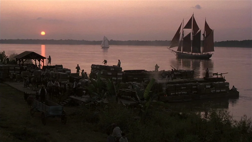 Interview With The Vampire movie still: establishing — A wide riverbank at sunset features wooden platforms laden with stacks of cotton bales.…