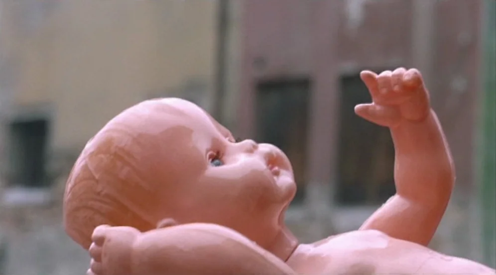Don’t Look Now (1973) movie still: closeup — A plastic baby doll sits in the foreground against a blurred cityscape background. The doll is…