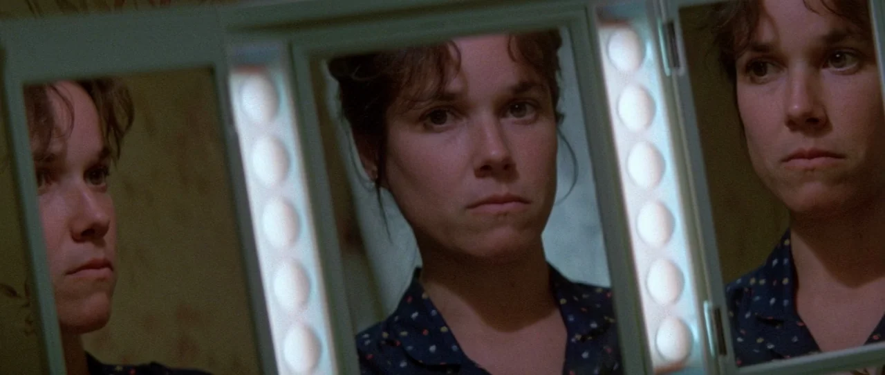 The Entity (1982) movie still: closeup — A woman looks into the central and side panels of a tri-fold vanity mirror. Her face is illuminated by…