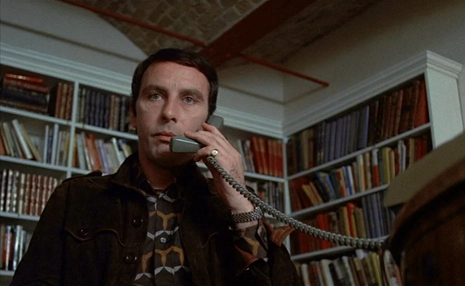 To The Devil a Daughter (1976) movie still: medium — A man stands in front of white bookshelves filled with numerous books. He is speaking into a…
