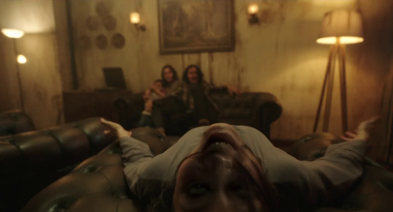 May The Devil Take You (2018) movie still: medium — A dimly lit room with peeling wallpaper and worn furniture in the background. A woman lies on…