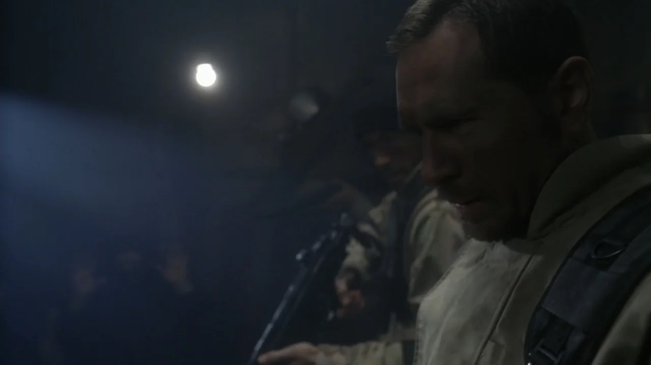 The Shepherd (2023) movie still: medium — A dark industrial setting features a man in tactical gear standing in shadow. He looks downward while…