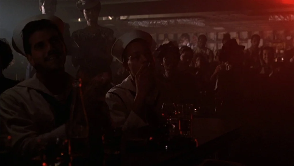 Bird (1988) movie still: medium — A dark, crowded room filled with patrons seated at tables in a dim environment. Two sailors in white caps sit in…