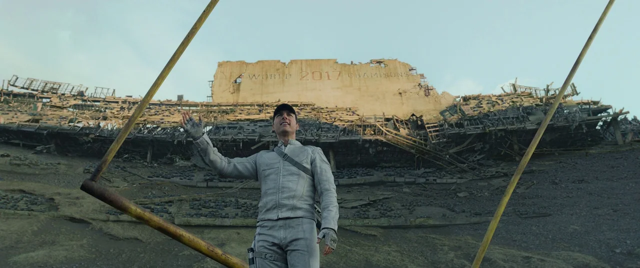 Oblivion (2013) movie still: fullbody — A vast, decaying sports arena lies in ruins under an overcast sky. A man in a grey suit gazes upward with…