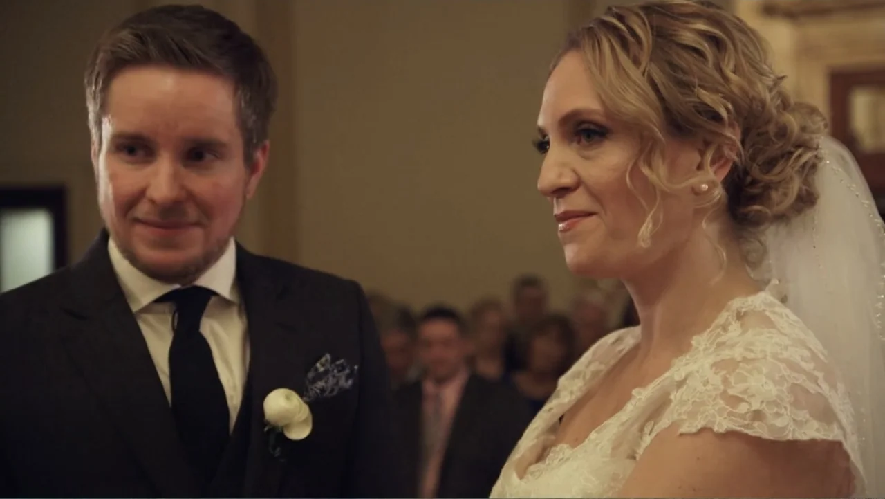 Suited (2016) movie still: medium — An indoor ceremony space filled with out-of-focus guests in the background. A bride and groom face each other…