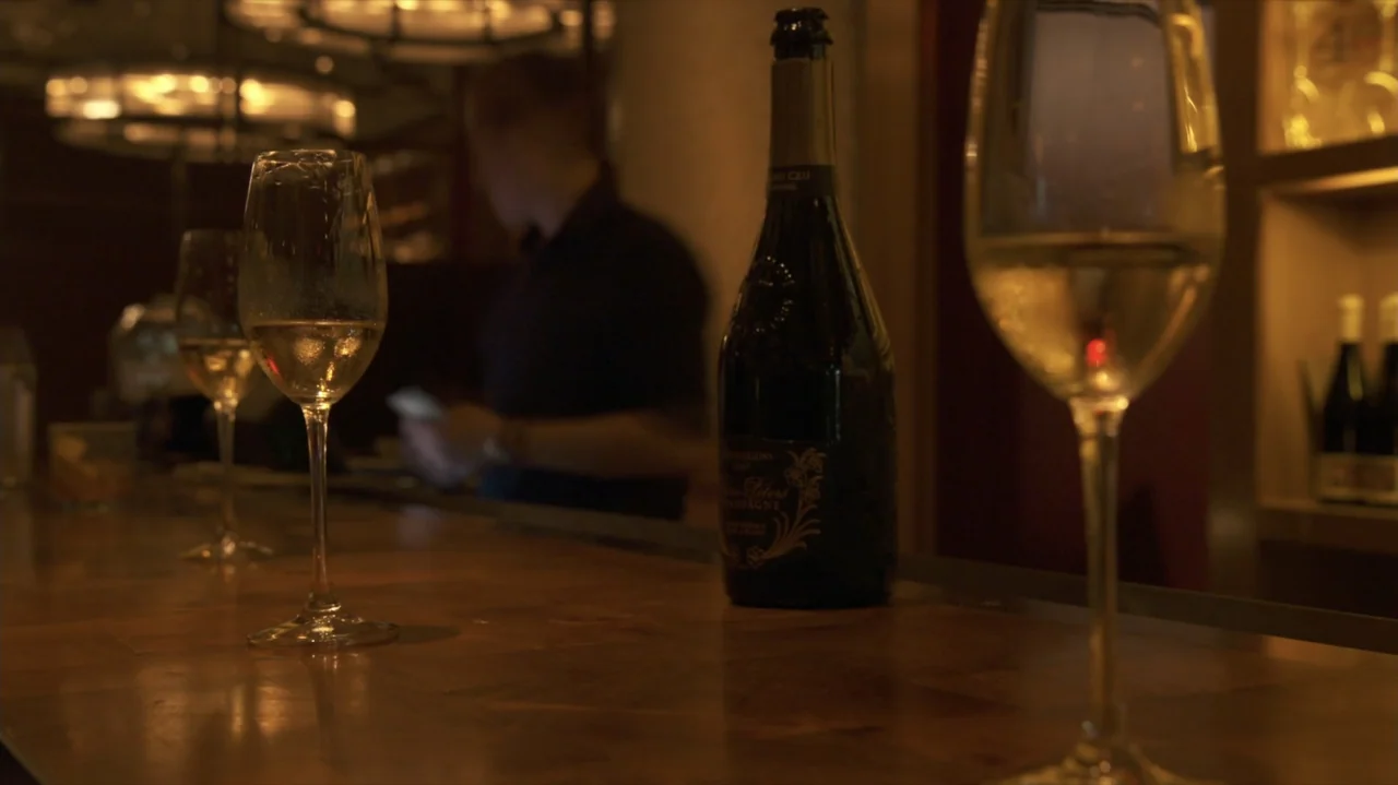 Somm: Into the Bottle (2015) movie still: medium — A wooden bar counter holds several wine glasses and a dark champagne bottle. The background is…