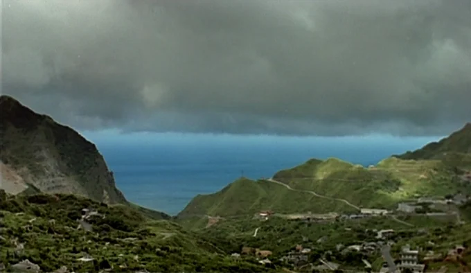 Dust In The Wind (1986) movie still: establishing — A wide view of a valley between green, sloped mountains leading to the ocean. A thin road winds…