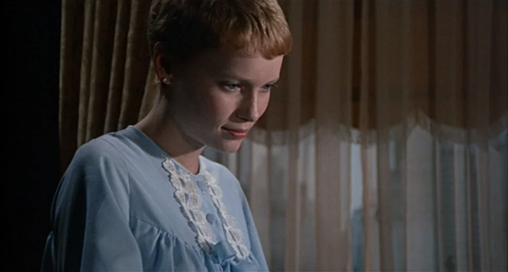 Rosemary’s Baby (1968) movie still: medium — A woman stands before a window covered by layered, semi-transparent beige curtains. She is looking down…