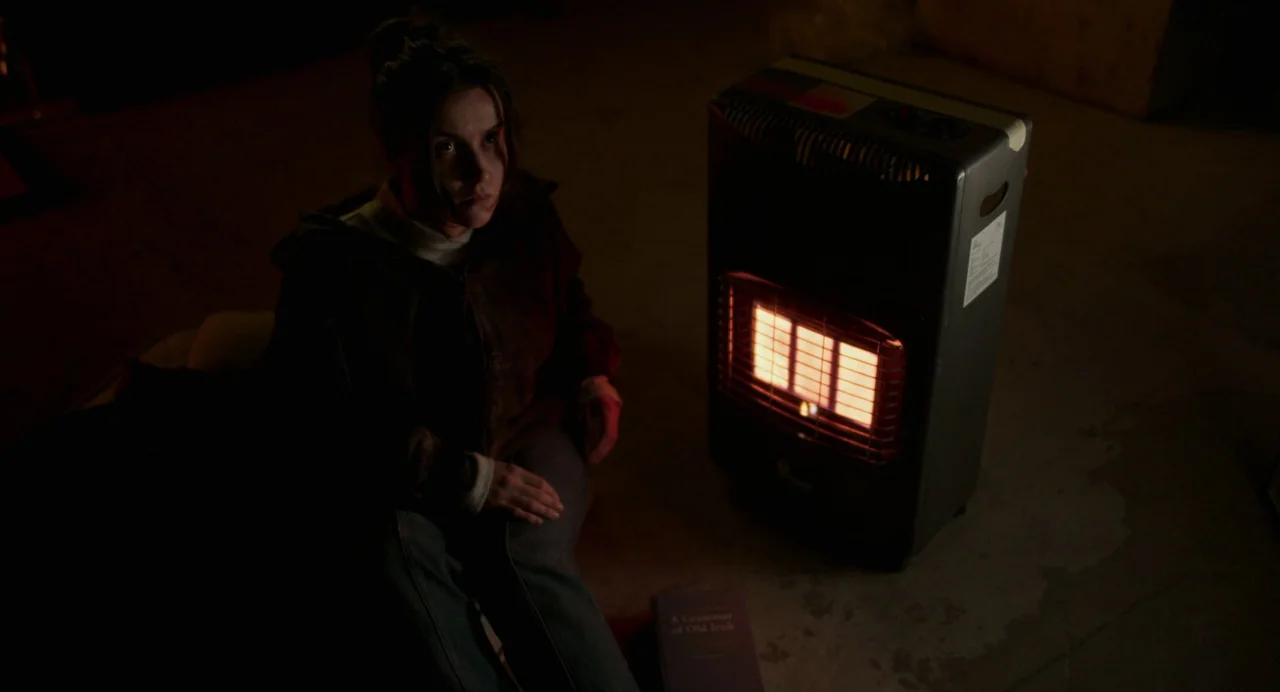 All You Need Is Death (2023) movie still: medium — A dark space with concrete floors is lit by a glowing portable gas heater. A young woman sits on…