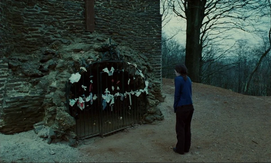 Hadewijch (2009) movie still: wide — An outdoor stone shrine with an iron gate stands against a wooded backdrop. A woman stands in front of the gate…