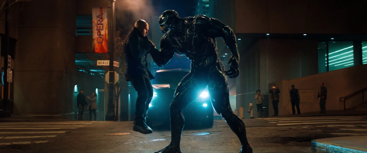 Venom (2018) movie still: fullbody — An urban intersection at night features a large alien creature lifting a man up. Pedestrians stand on the…