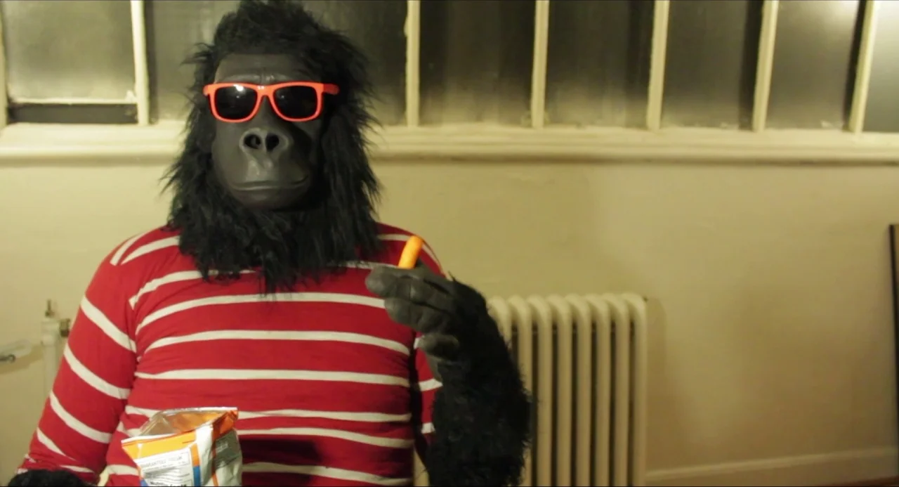 Like Me movie still: medium — An interior space with a beige wall, a white radiator, and a paneled window. A person wearing a gorilla mask and red…