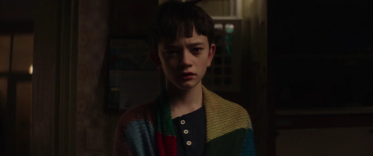 A Monster Calls (2016) movie still: A haunting, intimate portrait of a young boy draped in a colorful blanket, staring directly into the camera from…