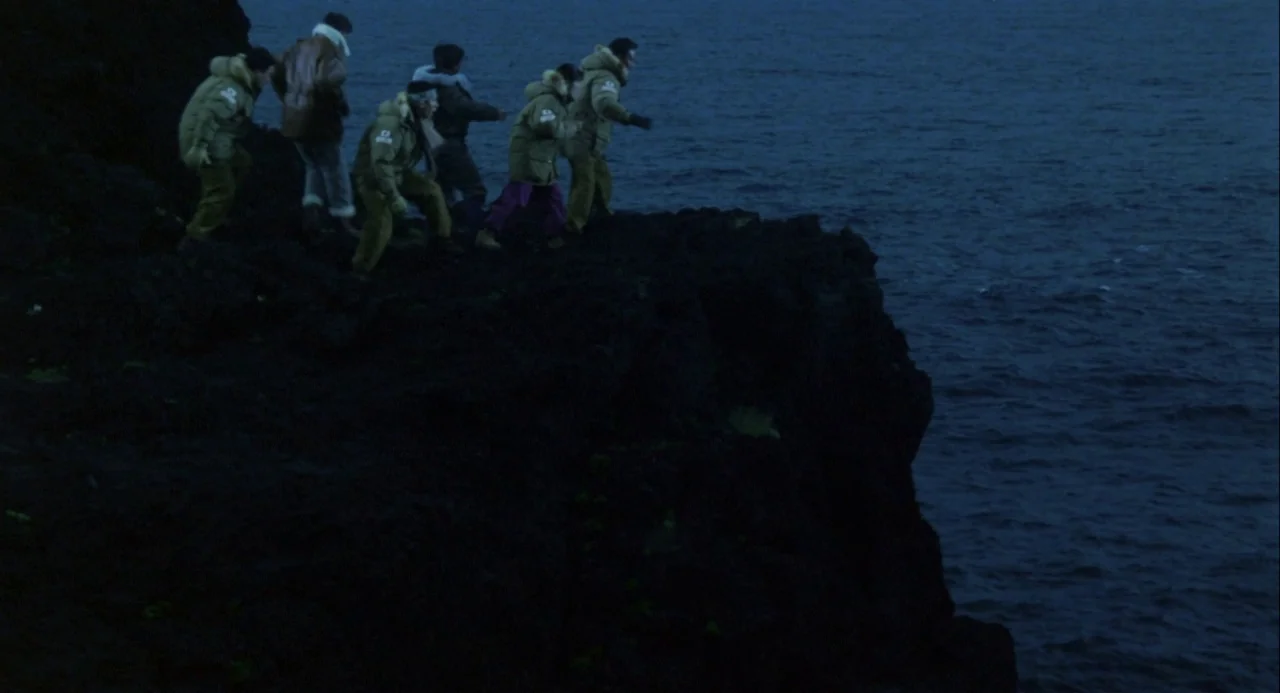 Godzilla vs. Mechagodzilla II (1993) movie still: wide — A group of people walks along the top of a dark, jagged coastal cliff. The deep blue sea…