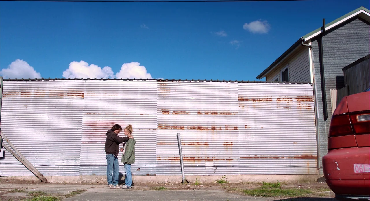 Killer Joe (2011) movie still: wide — A wide lot features a long corrugated metal wall with visible rust streaks. A man and a woman stand close to…
