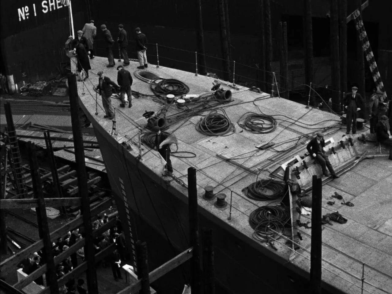 In Which We Serve (1942) movie still: wide — A group of workmen stand on the metal deck of a large vessel under construction. Coiled cables and…