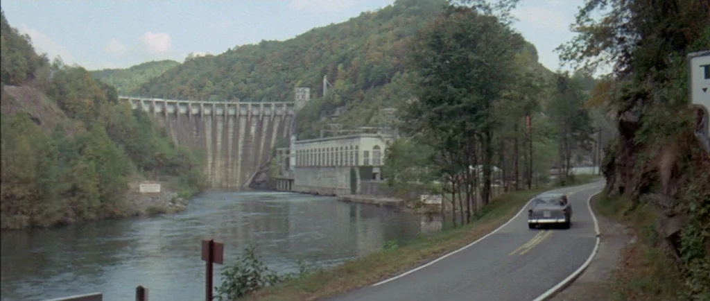 Two Lane Blacktop (1971) movie still: establishing — A concrete hydroelectric dam spans a river surrounded by forested hills during the day. A…
