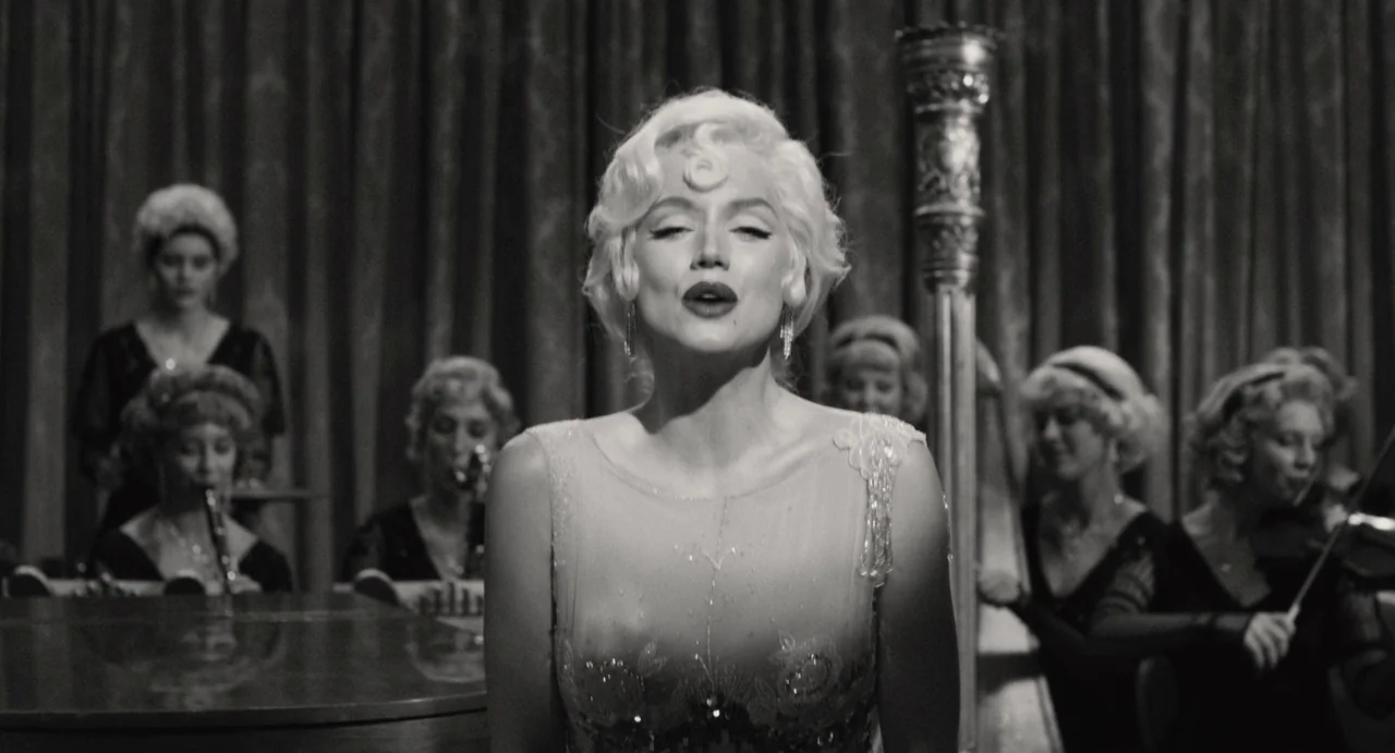 Blonde (2022) movie still: medium — A woman stands on a stage performing in front of a female musical ensemble. The background features heavy…