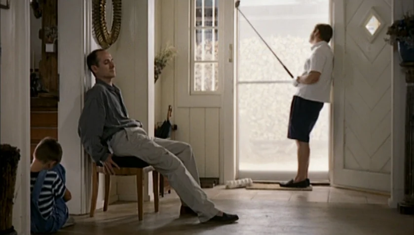 Funny Games (1997) movie still: wide — A sunlit residential hallway with light walls, tiled floors, and a screen door entry. A man sits on a chair,…
