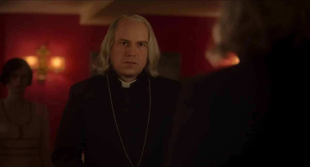 Men (2022) movie still: medium — A dimly lit room features dark red walls decorated with warm wall lamps. A middle-aged priest stands facing someone…