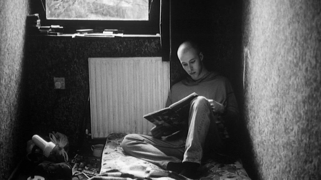 TwentyFourSeven (1997) movie still: medium — A cramped, dimly lit room with textured wallpaper features a single bed near a window. A young man sits…