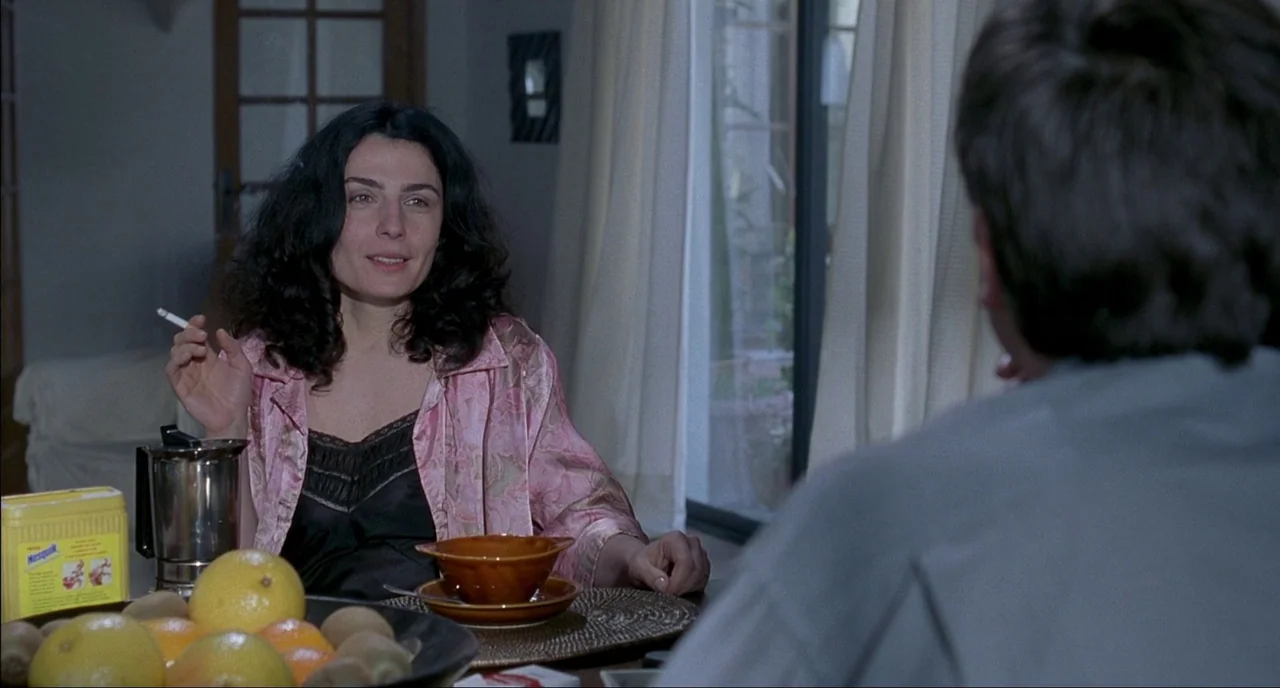Fat Girl (2001) movie still: medium — A woman sits at a dining table with fruit and coffee during the day. She smokes a cigarette while engaging in…