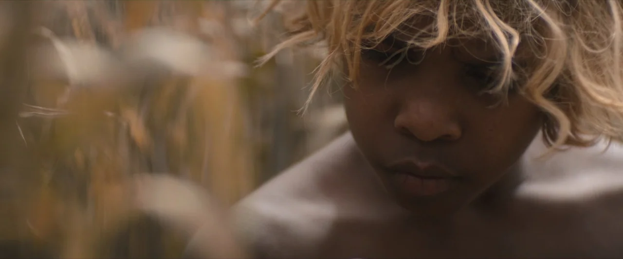 The New Boy (2023) movie still: closeup — An outdoor field filled with tall, dry grass and natural foliage. A young person with blonde hair looks…