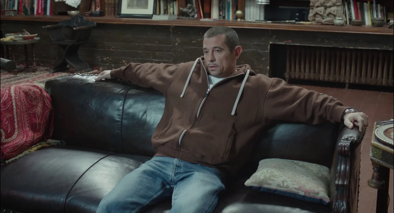 Abuse of Weakness (2013) movie still: medium — A man sits on a dark leather couch in a room with dark wood paneling. Wooden bookshelves filled with…