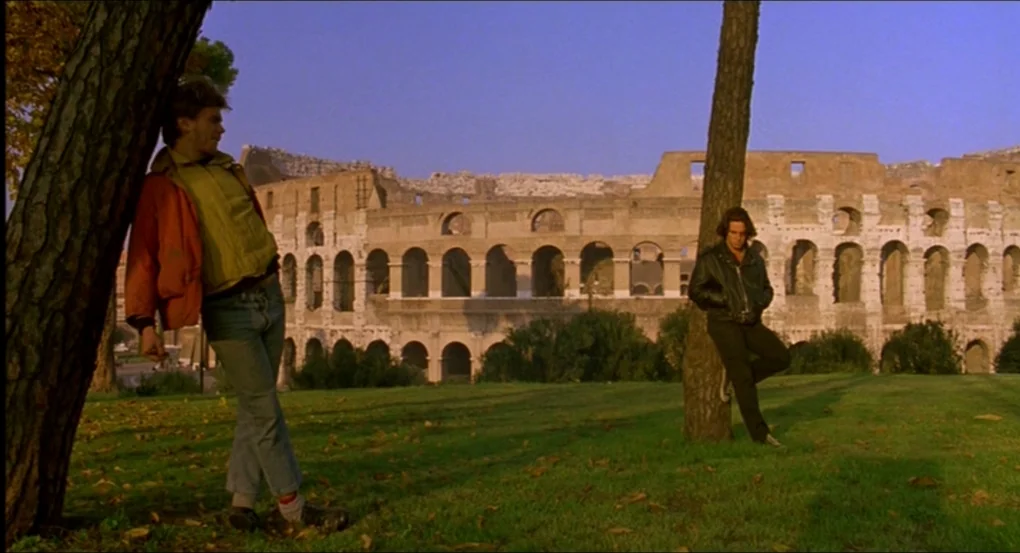 My Own Private Idaho (1991) movie still: wide — A grassy park area with scattered autumn leaves sits in front of the Colosseum. Two young men stand…