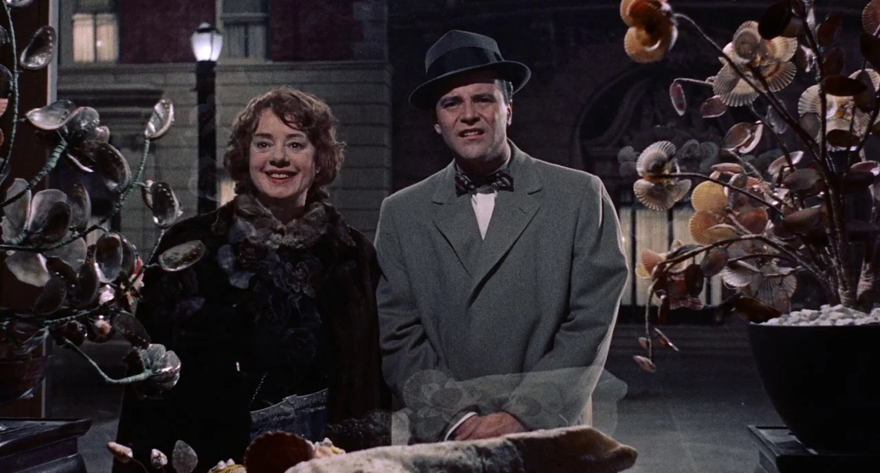 Bell Book and Candle (1958) movie still: medium — An urban setting at night features two people standing behind decorative shell displays. The woman…