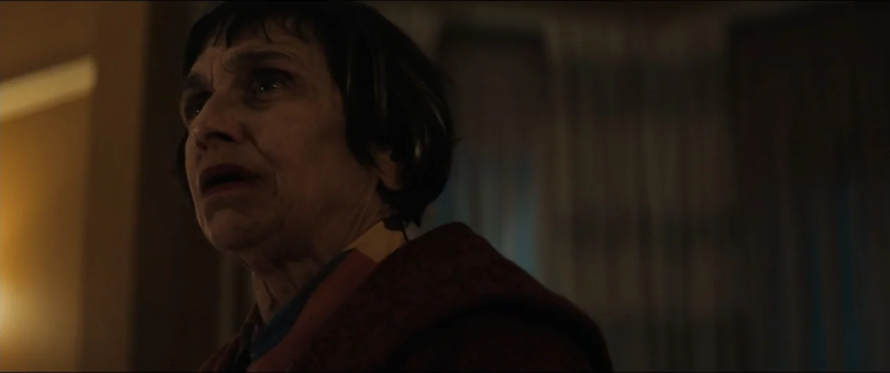 Vicious (2025) movie still: closeup — A dimly lit indoor space features a woman in a dark coat standing near curtains. The woman looks toward the…