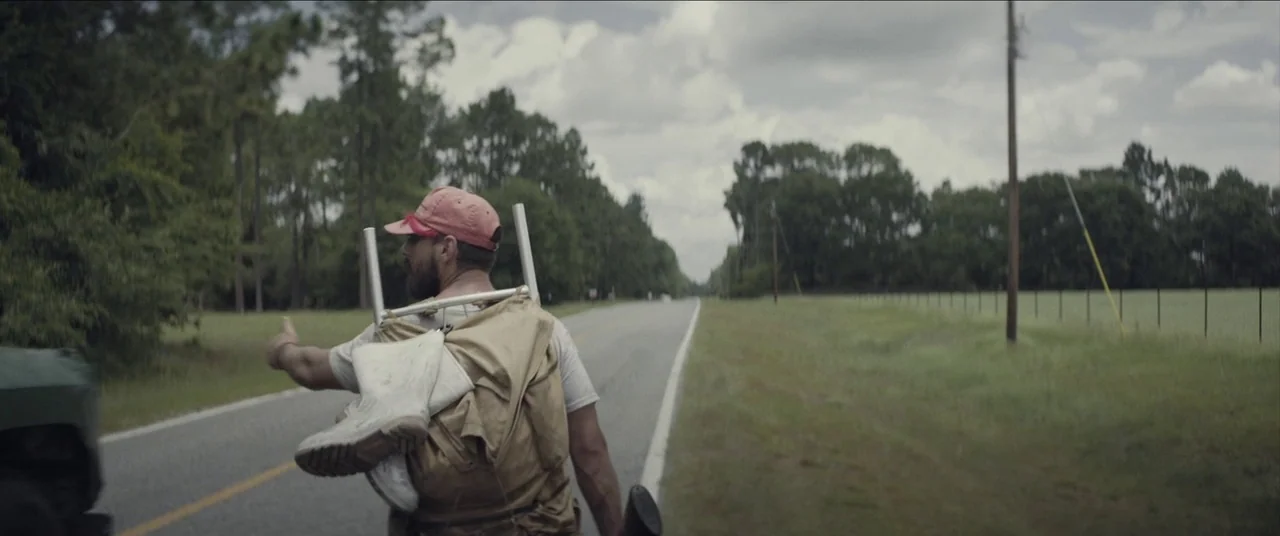 The Peanut Butter Falcon (2019) movie still: medium — A man stands on the edge of a paved rural road bordered by trees. He wears a large backpack…
