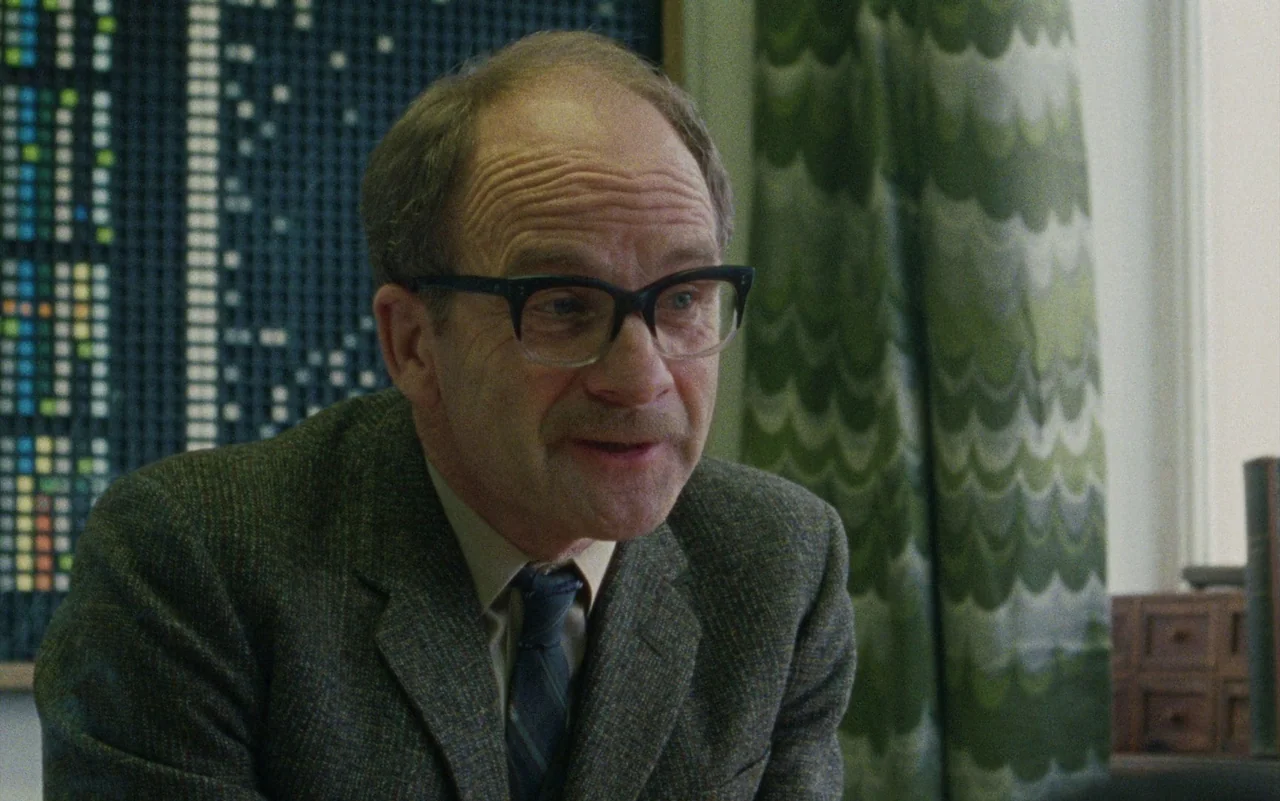 Education (2020) movie still: closeup — A man sits in an office featuring a grid-patterned wall and patterned green curtains. The man speaks while…