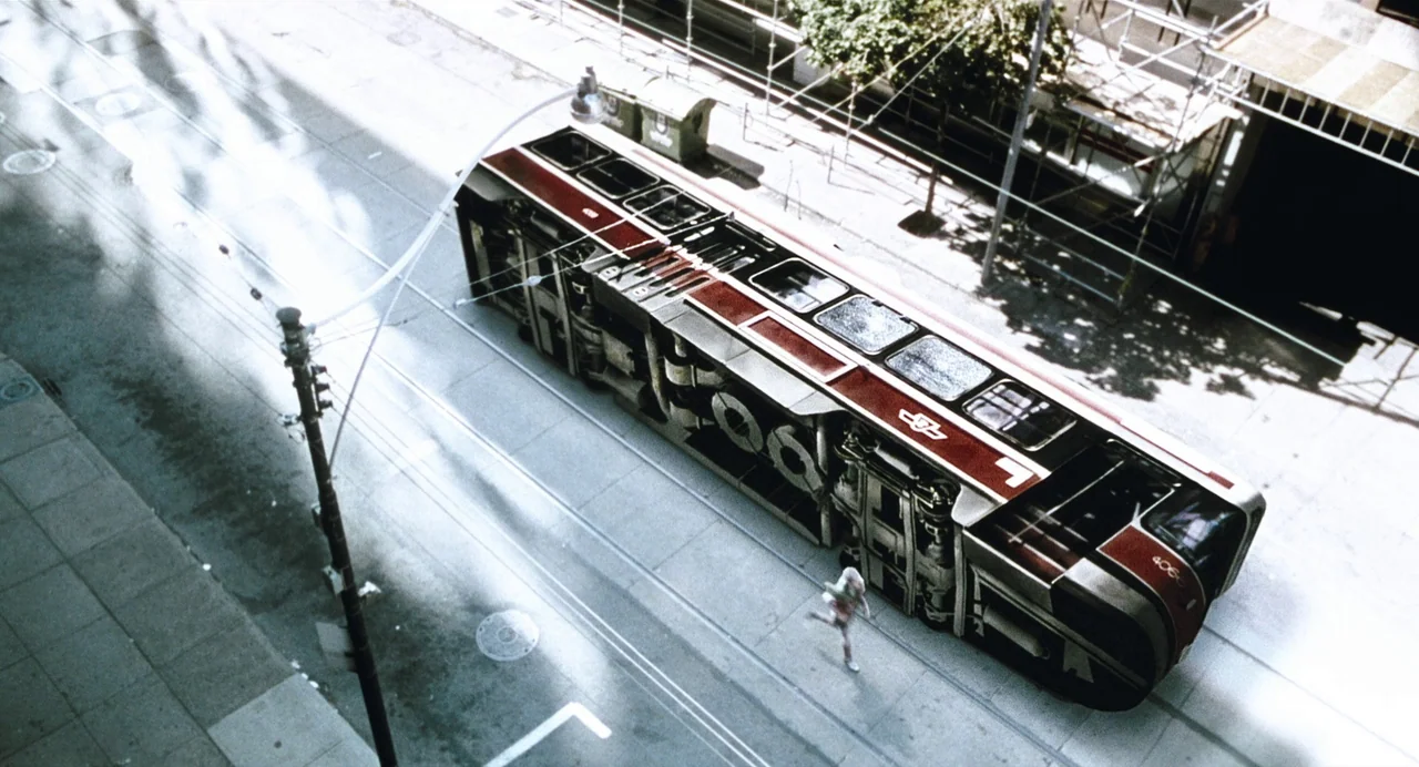 Last Night (1998) movie still: wide — A metropolitan street features an overturned multi-car public transit tram on its side. A small person in…