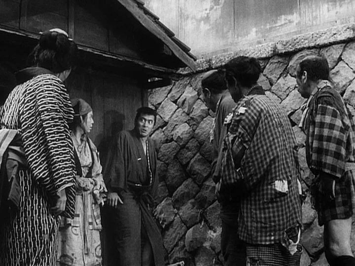 The Lower Depths (1957) movie still: fullbody — A group of people in traditional Japanese clothing stand near a rough stone wall. A man in a dark…