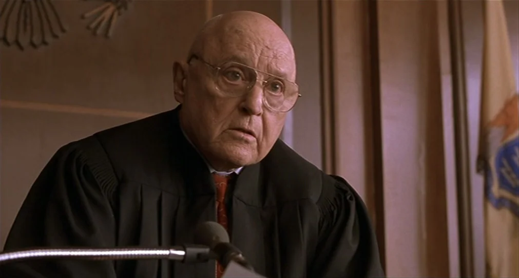 The Hurricane (1999) movie still: medium — A courtroom setting featuring wood-paneled walls and a vertical state flag. An elderly male judge sits at…