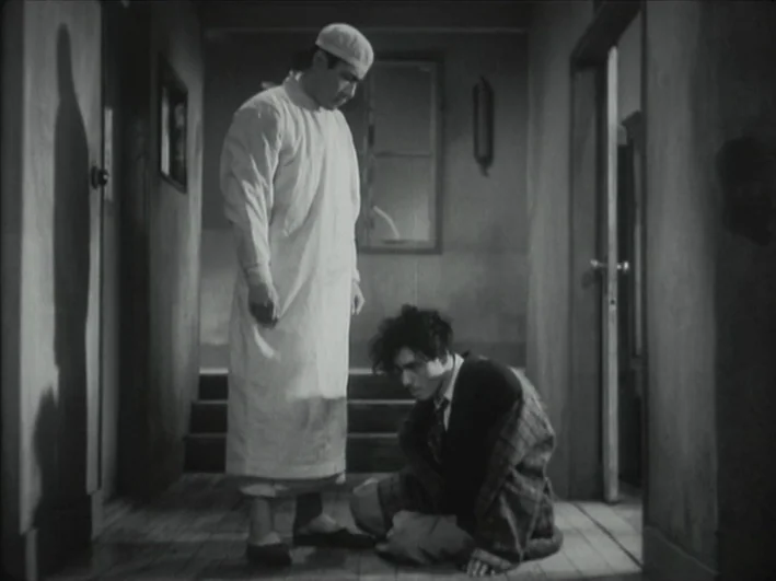 The Quiet Duel (1949) movie still: fullbody — A dimly lit narrow hallway with wood floors and doorways on either side. A man in medical clothes…