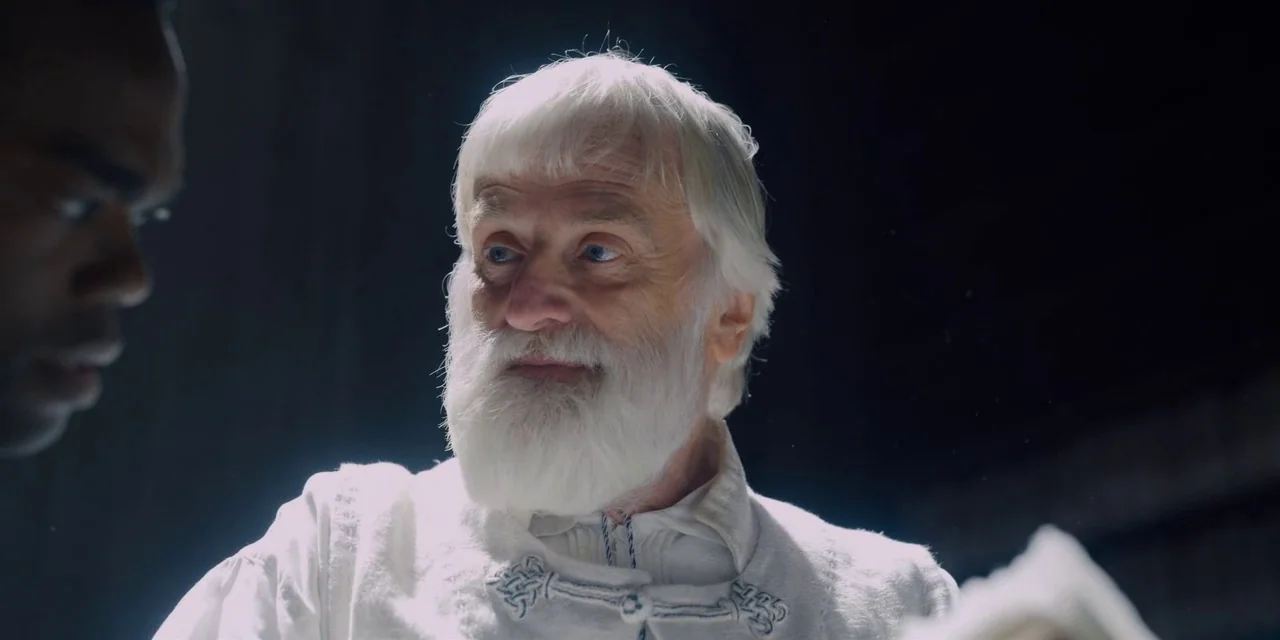 Midsommar (2019) movie still: closeup — The setting is a dark interior space with minimal architectural detail visible. An elderly white-haired man…