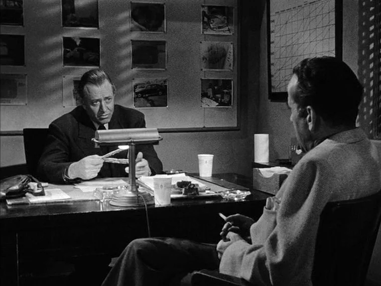 In a Lonely Place (1950) movie still: medium — A dim office wall is covered with a grid of photographic prints and charts. A man in a suit sits at a…