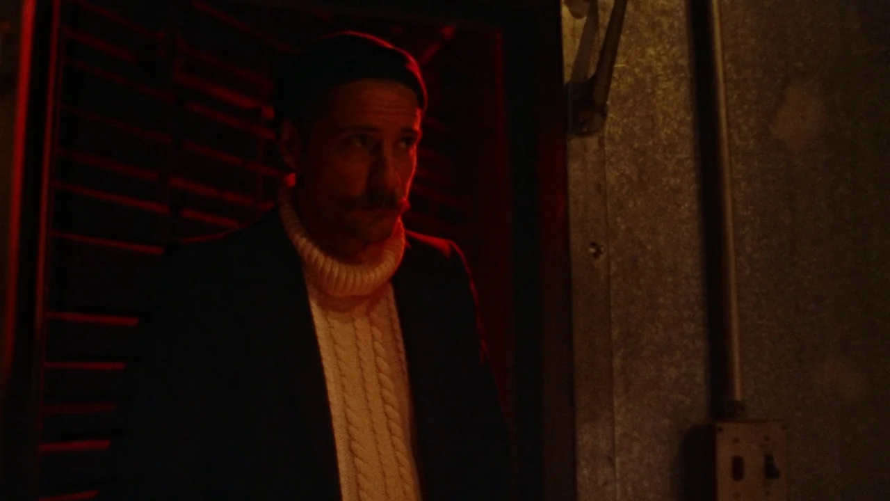 Strawberry Mansion (2021) movie still: medium — A dimly lit industrial storage area with metallic walls and a red-lit doorway. A man in a sweater…