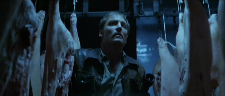 Roadgames (1981) movie still: medium — A dark meat locker filled with multiple raw animal carcasses hanging from metal hooks. A man stands amidst…