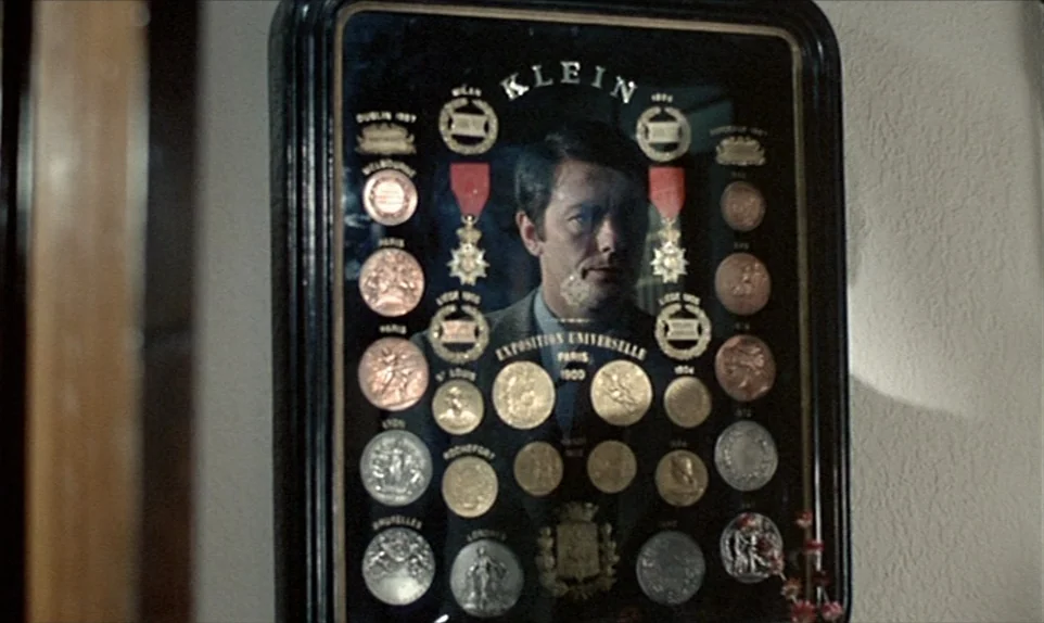 Mr. Klein (1976) movie still: closeup — A man's face is visible as a reflection on a glass-encased collection of medals. The wooden display board is…