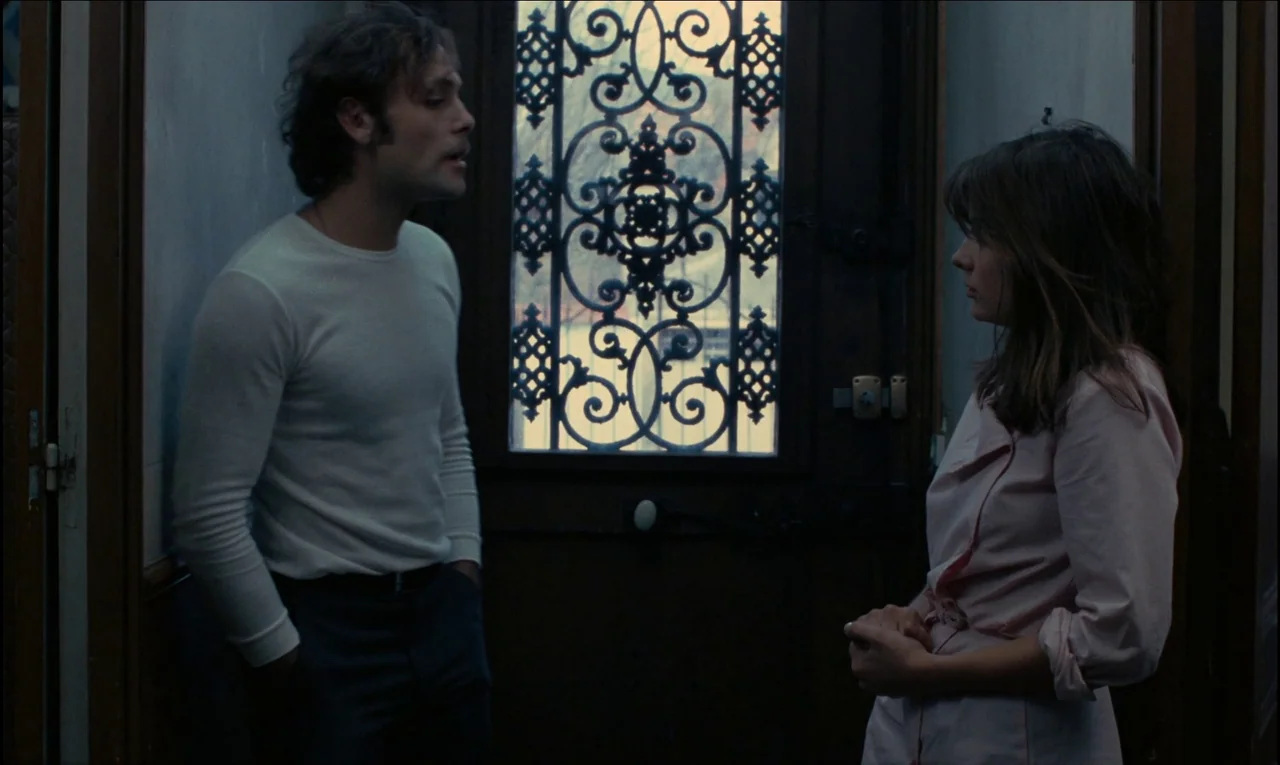 Serie Noire (1979) movie still: medium — A dimly lit residential entryway features a dark wooden door with elaborate iron grillwork. A man in a…