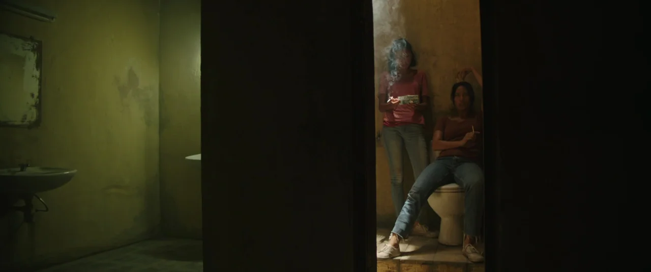 Impetigore (2019) movie still: wide — A cramped, dilapidated room with peeling yellow paint and a visible ceramic sink unit. Two young women occupy…
