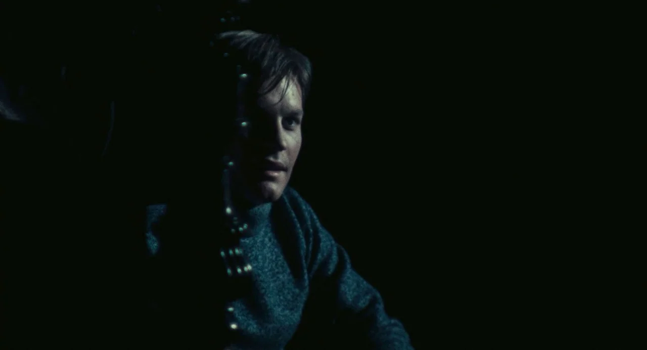 The Damned (1969) movie still: medium — A man sits in an extremely dark environment with minimal visible surroundings. He turns his head while…